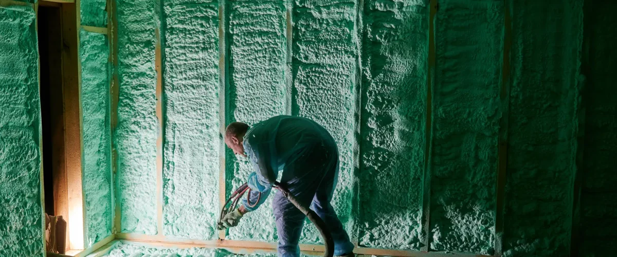 Professional Spray Foam Insulation — Attic Application Technician spraying closed-cell foam insulation on attic walls and ceiling in a wood-frame house.
